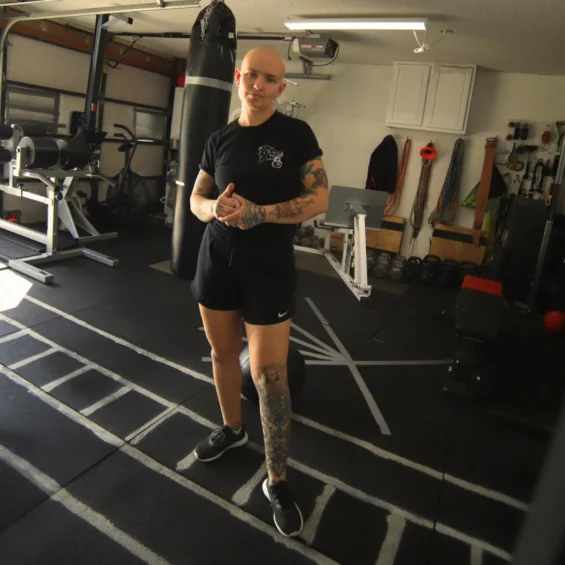 Annie Spence, local young entrepreneur with a shaved head, tattoos and black gym clothes, stands in front of gym equipment.