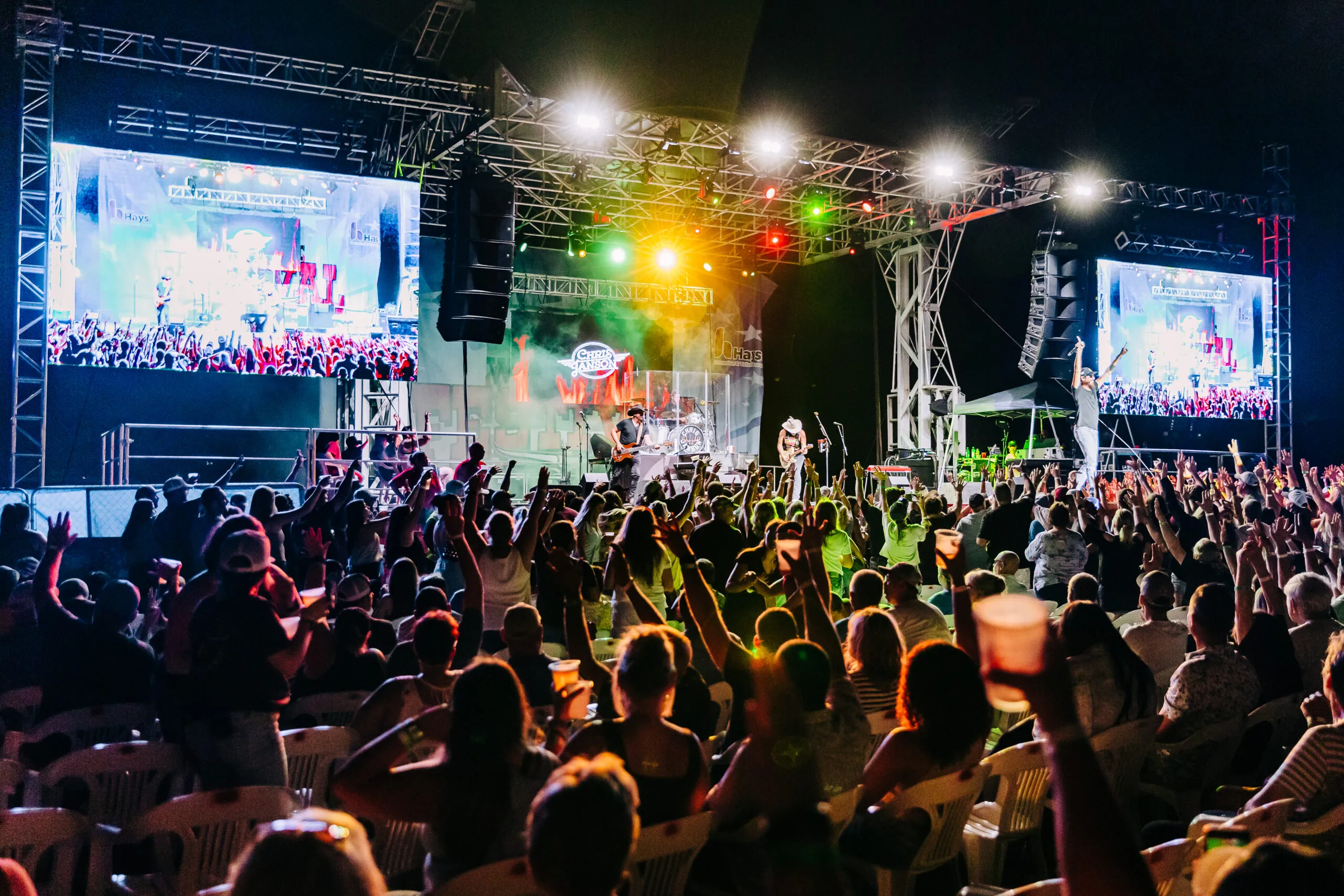 A large crowd in front of an outdoor stage at night with a band and colorful lights.