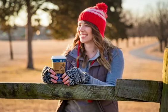 Woman in a hat and gloves holding a cup of coffee in a field in the early morning.