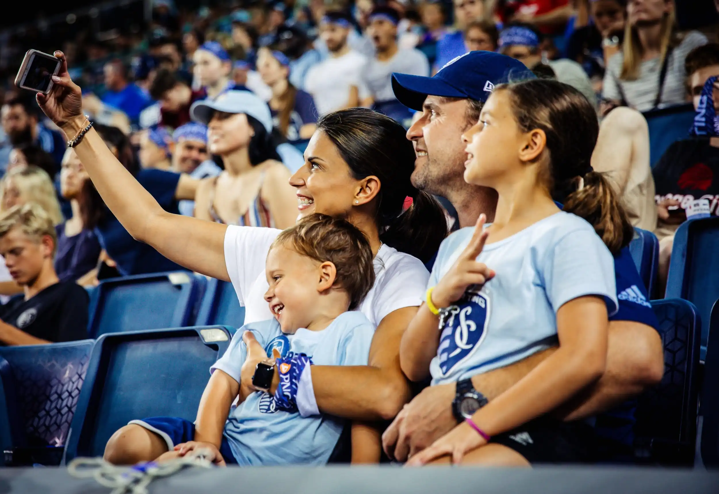 Parents and two children smile for a selfie in the stands at a Sporting KC MLS game.